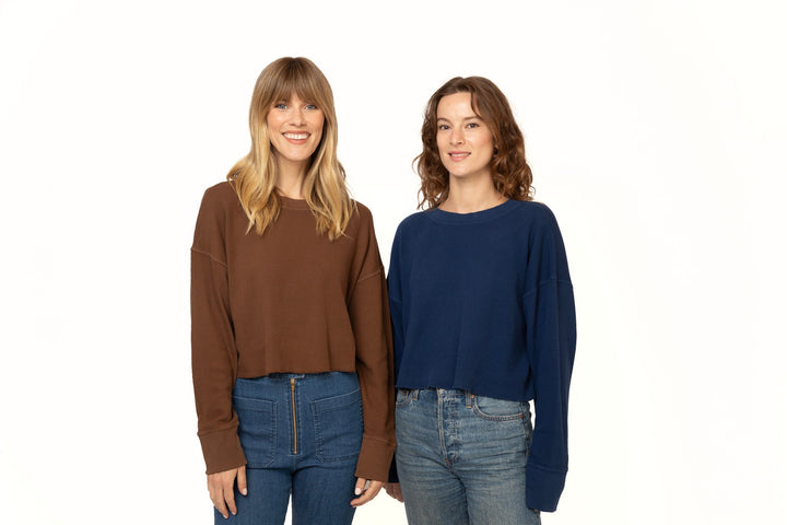 Two women wearing brown and blue sweaters with jeans on a white background